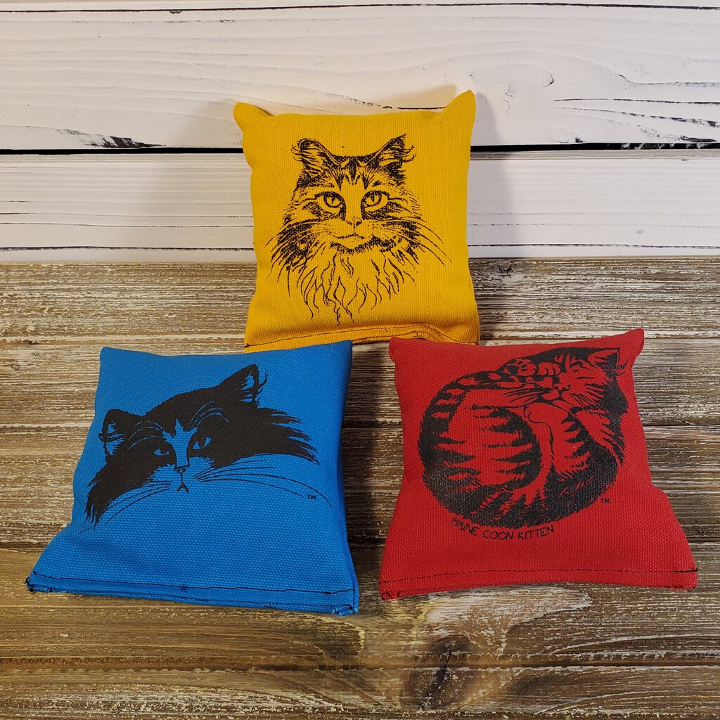 Three catnip sacks in red, blue, and yellow colors, each with a printed cat face on it, placed on a wooden surface.