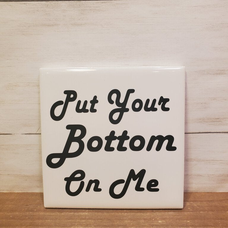 Tile with text 'Put Your Bottom On Me' on a wooden surface