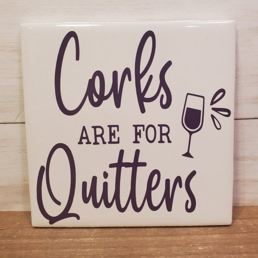 A ceramic coaster with the text 'Corks Are For Quitters' printed on it, along with an illustration of a wine glass.