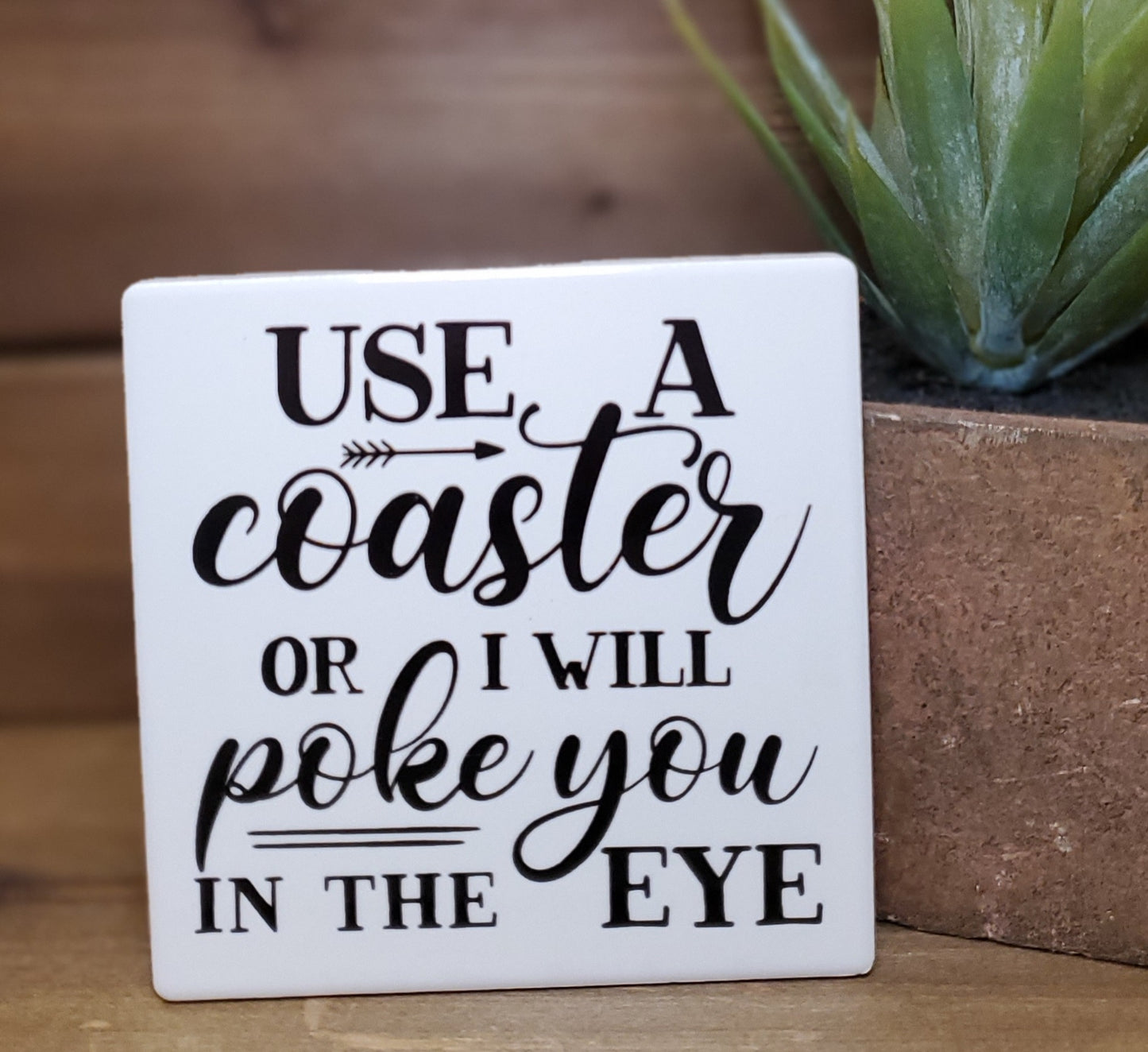 White coaster with humorous text on a wooden surface with a plant in the background