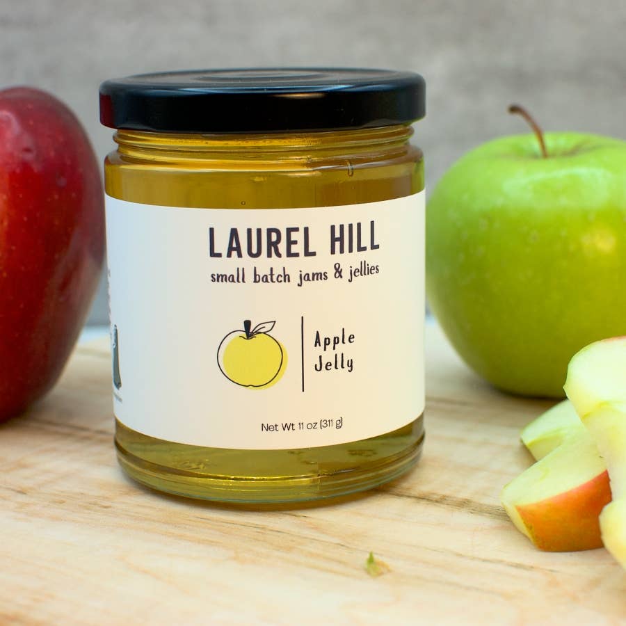 A jar of Laurel Hill Apple Jelly sits on a wooden cutting board with a red apple, a green apple, and apple slices around it. The jar has a white label with black text and a black lid.