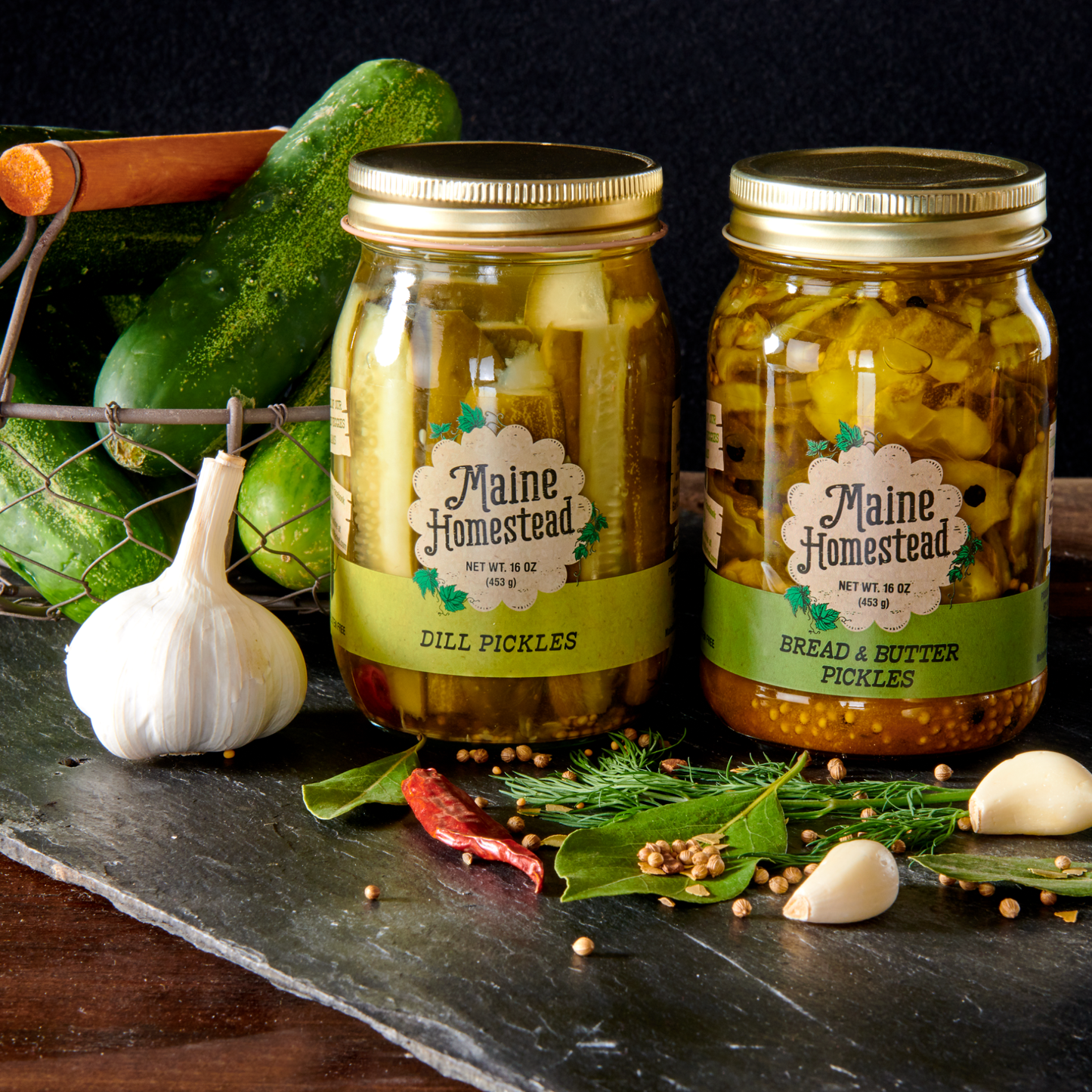 Two jars of pickles with 'Maine Homestead' labels on a dark surface with cucumbers and garlic.