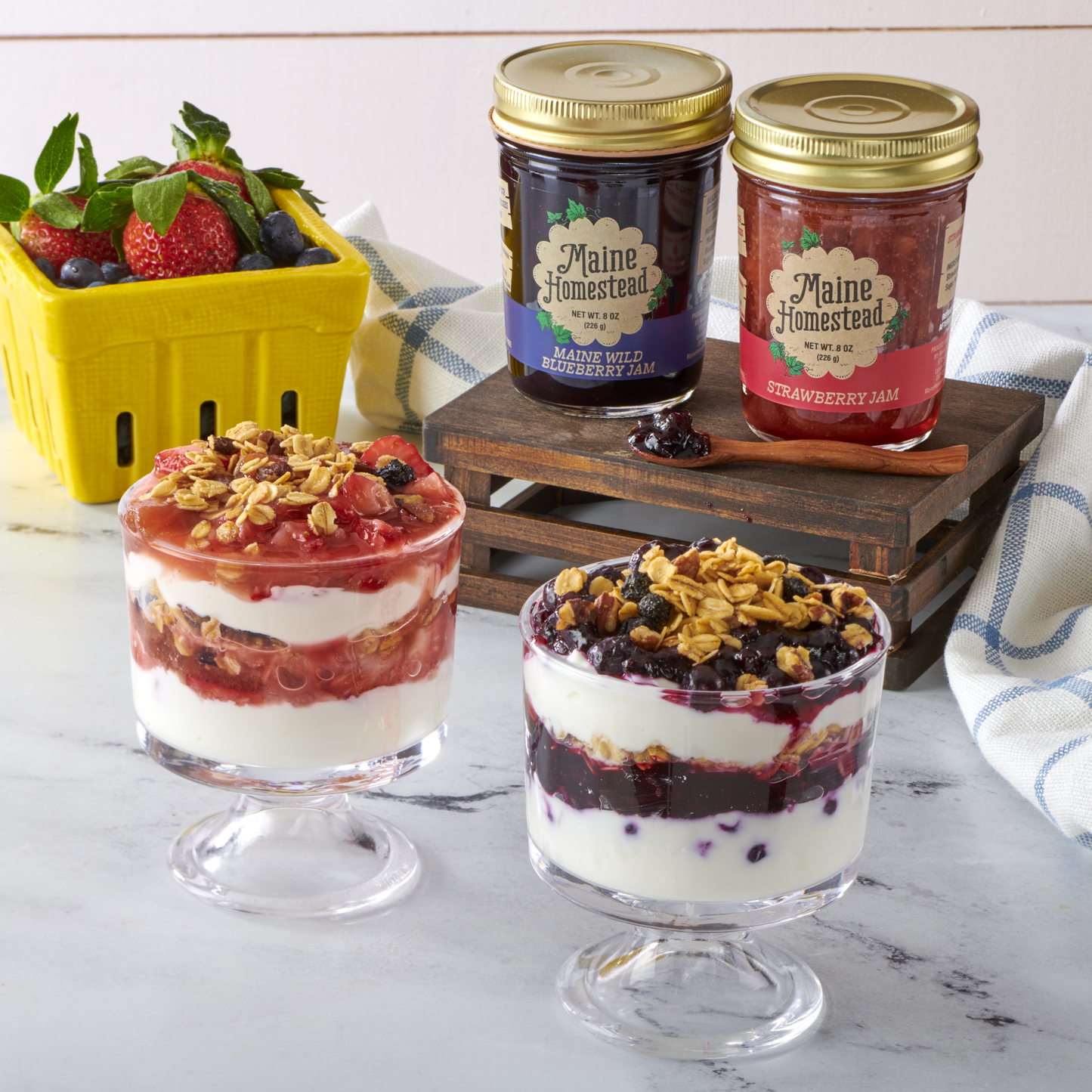 Two glass dessert dishes with layered yogurt and fruit, accompanied by jars of Maine Homestead jam and a container of strawberries.