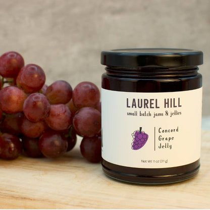 A jar of Laurel Hill Concord Grape Jelly sits on a light wooden surface next to a bunch of red grapes. The jar has a black lid and a white label with a purple grape illustration.