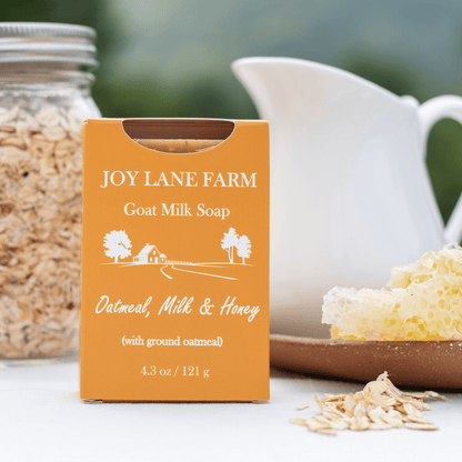 A box of Joy Lane Farm Goat Milk Soap stands on a tabletop with oatmeal flakes, a jar of oats, a white pitcher, and honeycomb in the background. The box is orange with white text reading 'Joy Lane Farm Goat Milk Soap' and 'Oatmeal, Milk & Honey (with ground oatmeal)'.