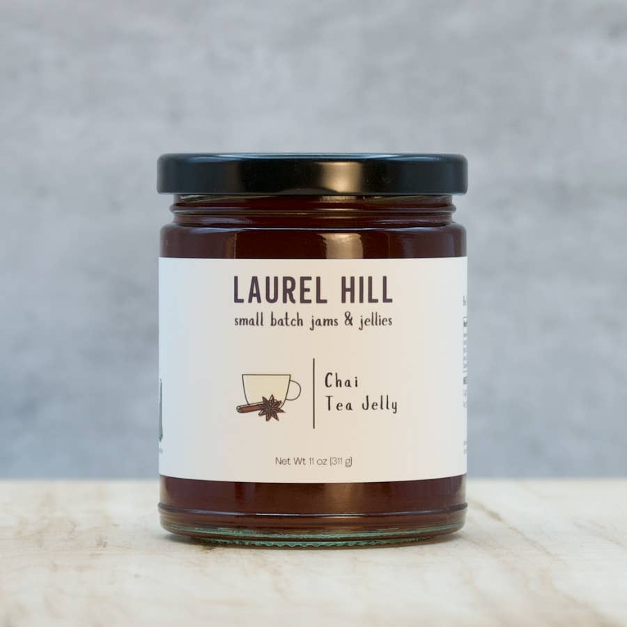 A jar of Laurel Hill Chai Tea Jelly on a light marble surface with a textured gray background. The product is in a clear glass jar filled with dark brown jelly, featuring a cream-colored label with black text and a black lid.