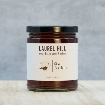 A jar of Laurel Hill Chai Tea Jelly on a light marble surface with a textured gray background. The product is in a clear glass jar filled with dark brown jelly, featuring a cream-colored label with black text and a black lid.