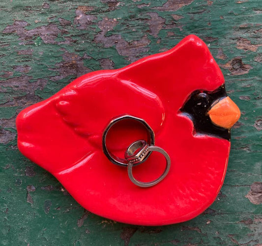 A red ceramic bird-shaped ring holder with a black beak and orange nose, holding two silver rings, set against a distressed green wooden background.