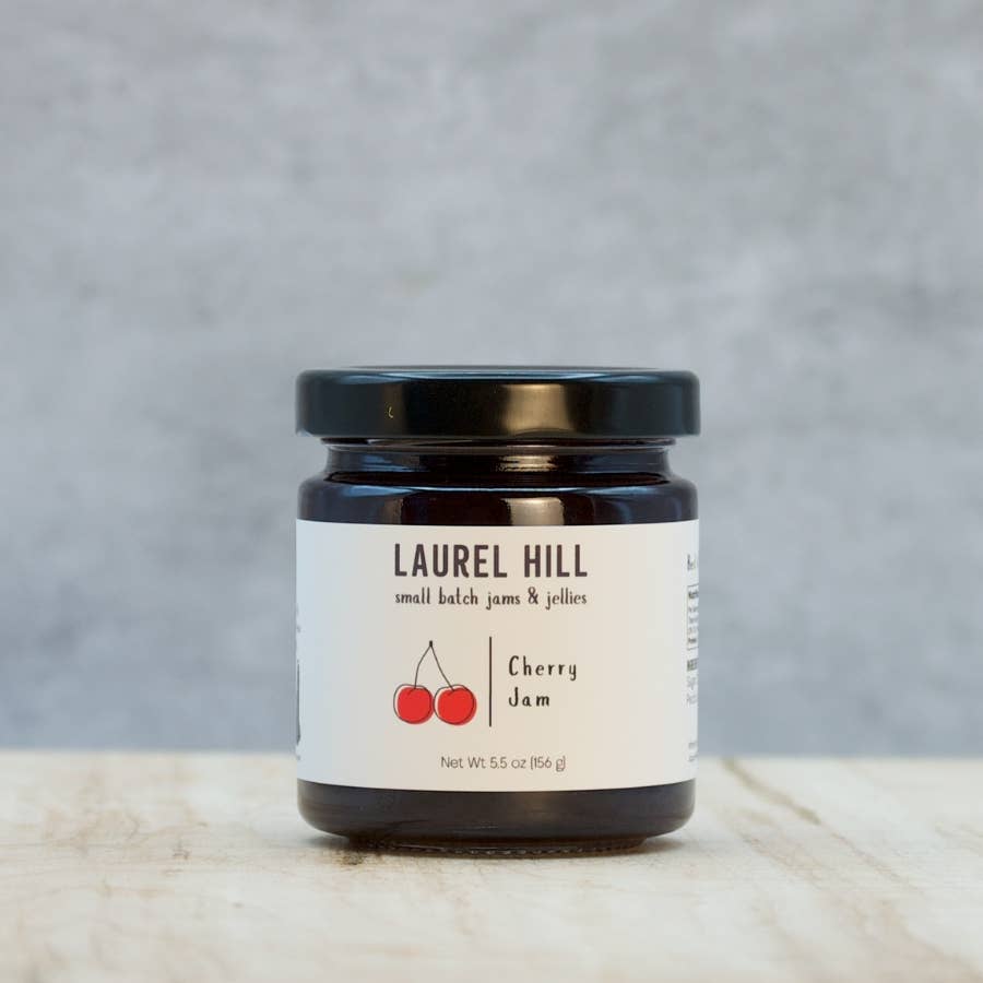A jar of Laurel Hill Cherry Jam on a light wooden surface with a textured gray background. The product is in a dark glass jar with a black lid and a white label featuring the brand name and red cherry illustration.