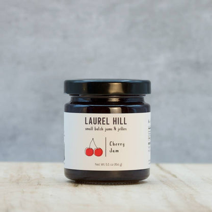 A jar of Laurel Hill Cherry Jam on a light wooden surface with a textured gray background. The product is in a dark glass jar with a black lid and a white label featuring the brand name and red cherry illustration.