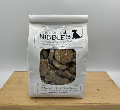 A white stand-up pouch of North i Woods Nibbles All Natural Small Dog Treats, featuring a clear window showing round brown cheddar-flavored treats inside, set on a wooden board against a neutral gray background.