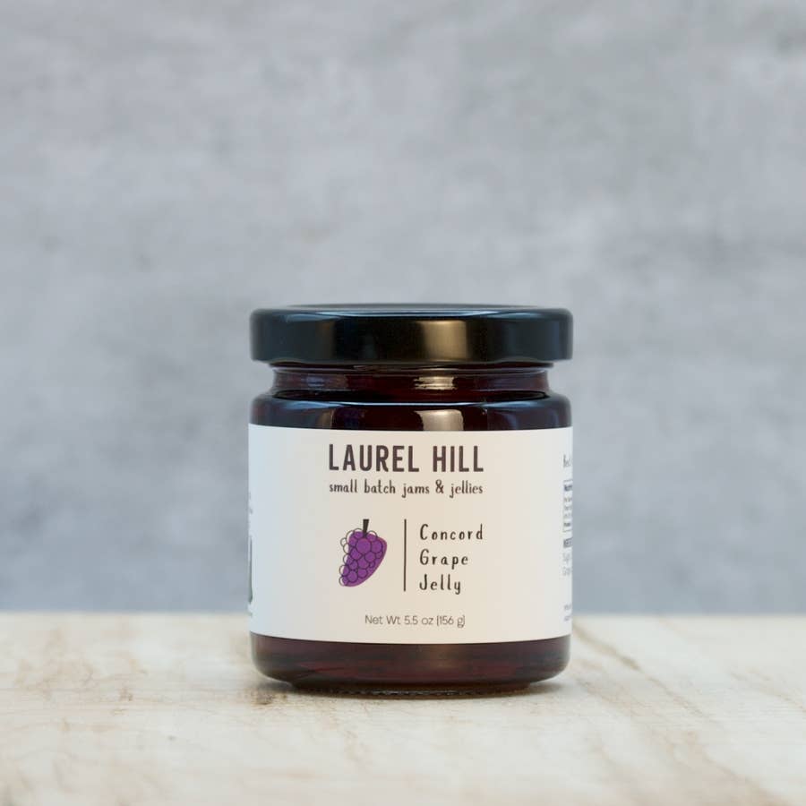 A jar of Laurel Hill small batch jam & jellies, Concord Grape Jelly, on a light marble surface with a textured gray background.