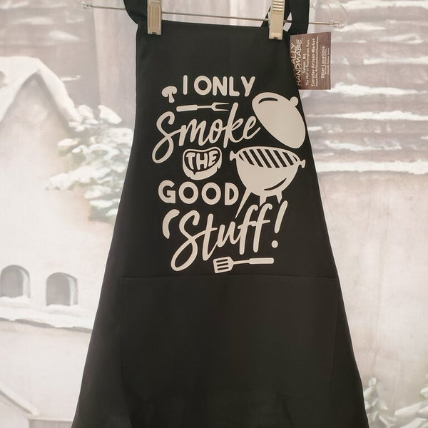 A black apron with white text 'I ONLY SMOKES THE GOOD STUFF!' and an illustration of a grill printed on it, displayed against a rustic background.