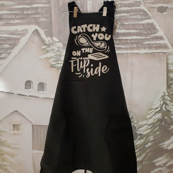 A black apron with white text 'CATCH YOU ON THE FLIP SIDE' printed on the front, displayed against a wintry backdrop with snow-covered houses.
