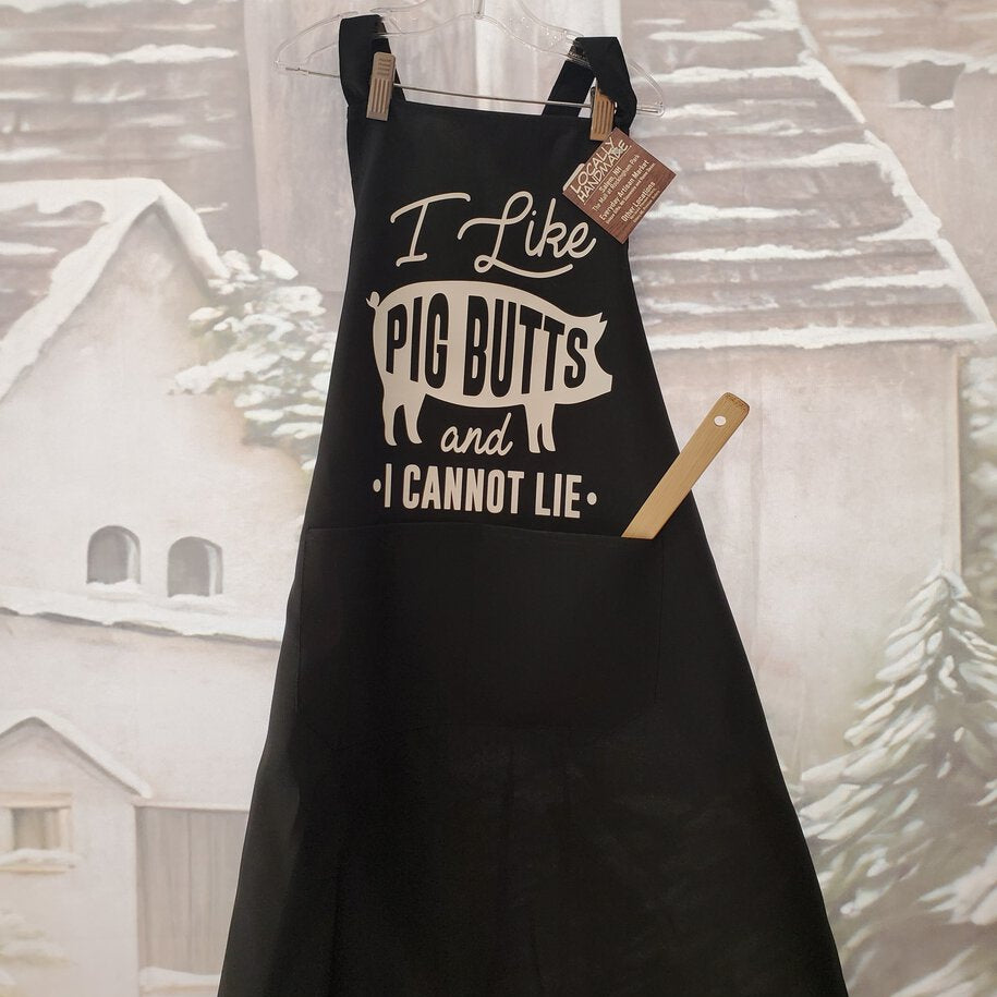 A black apron with the text 'I Like Pig Butts and I Cannot Lie' printed on it, displayed against a festive background with Christmas trees.