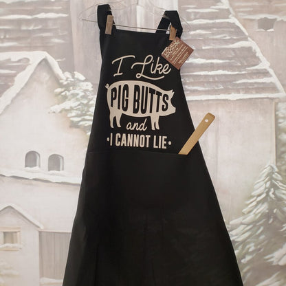 A black apron with the text 'I Like Pig Butts and I Cannot Lie' printed on it, displayed against a festive background with Christmas trees.