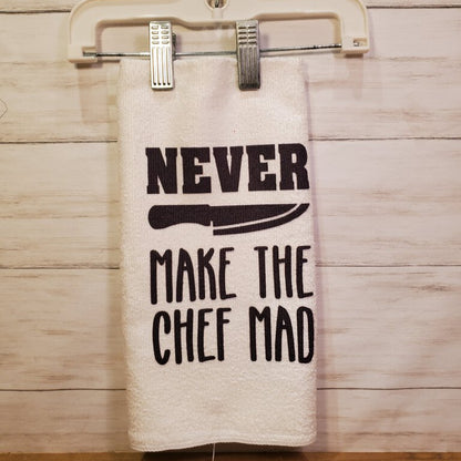 A white kitchen towel with black text and a graphic of a knife, hanging on a rod against a wooden backdrop. The text reads 'NEVER MAKE THE CHEF MAD'.
