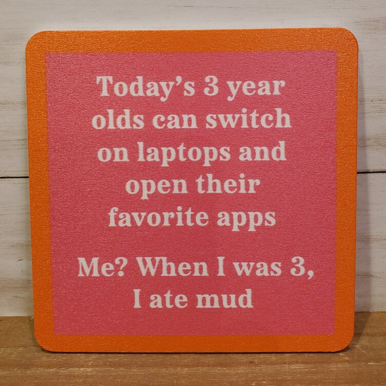 A square coaster with orange edges and a red center, featuring white text that reads: 'Today's 3 year olds can switch on laptops and open their favorite apps. Me? When I was 3, I ate mud.