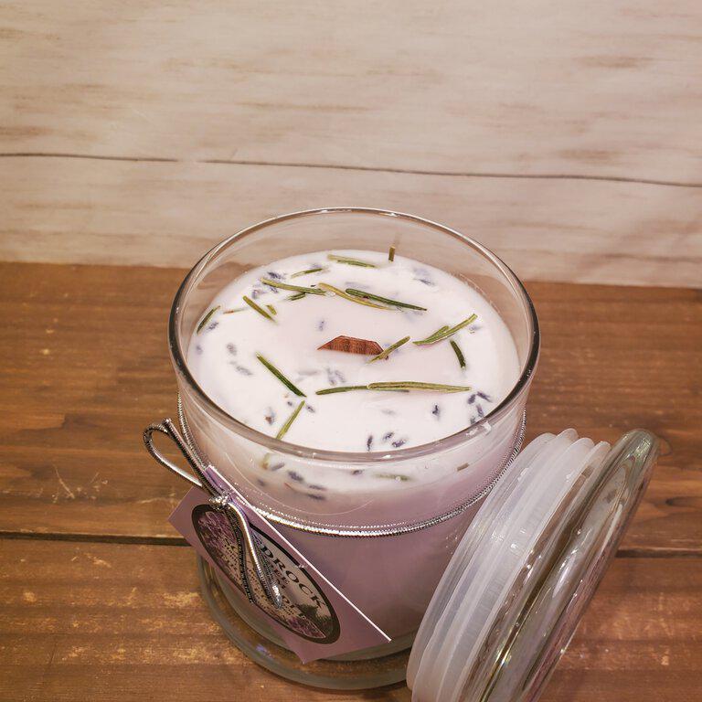 A lavender-colored soy wax candle inside a clear glass jar with a decorative lid, set on a wooden surface. The candle is adorned with lavender sprigs and has a white, ribbon-like decoration around the wick.