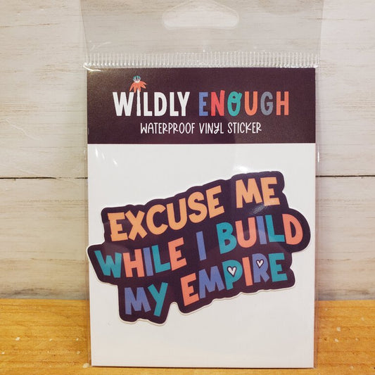 A sticker with the phrase 'Excuse Me While I Build My Empire' printed in multicolored letters, packaged on a card.