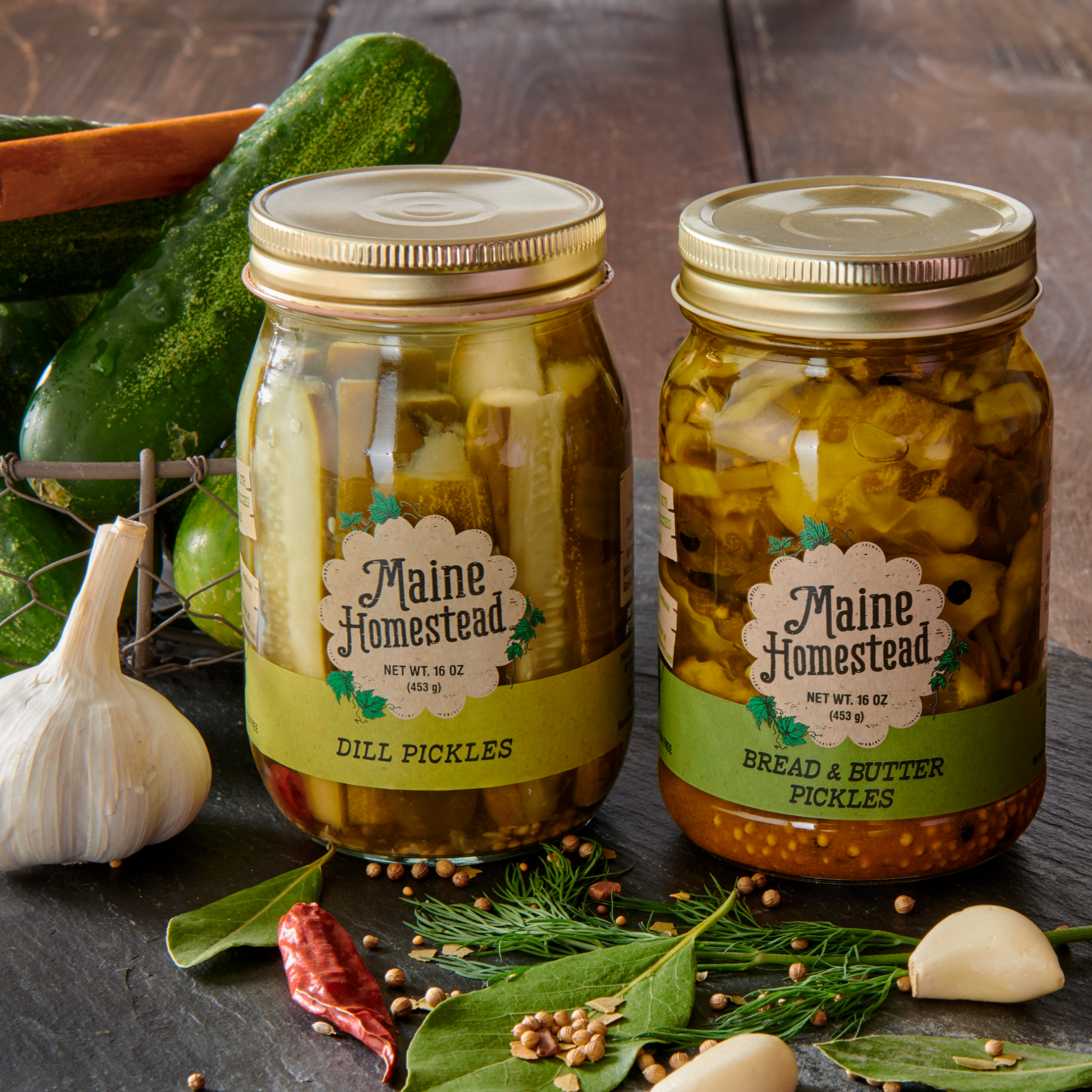 Two jars of pickles from Maine Homestead on a wooden surface with cucumbers and garlic.