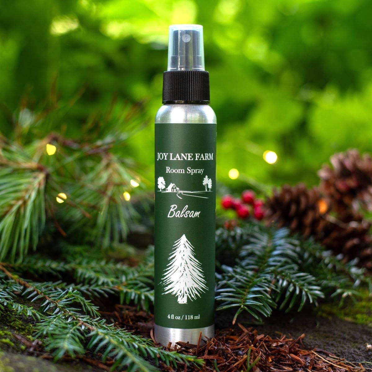 A bottle of Joy Lane Farm Balsam Room Spray stands outdoors on evergreen branches with pinecones and soft green bokeh in the background. The bottle is dark green with white text and a white tree illustration, and it has a black spray top.