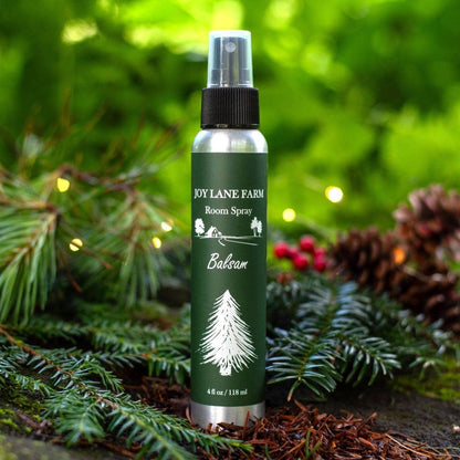 A bottle of Joy Lane Farm Balsam Room Spray stands outdoors on evergreen branches with pinecones and soft green bokeh in the background. The bottle is dark green with white text and a white tree illustration, and it has a black spray top.