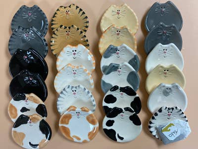 A collection of ceramic cat-shaped plates or dishes arranged in three columns on a light beige background. The plates feature various cat breeds and patterns, including black and white, calico, tabby, and spotted designs, with some plates showing printed cat faces.