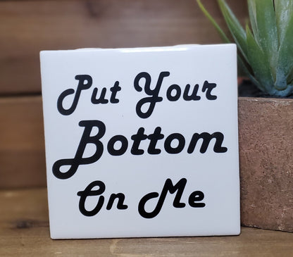 White square tile with black text 'Put Your Bottom On Me' next to a plant on a wooden surface.