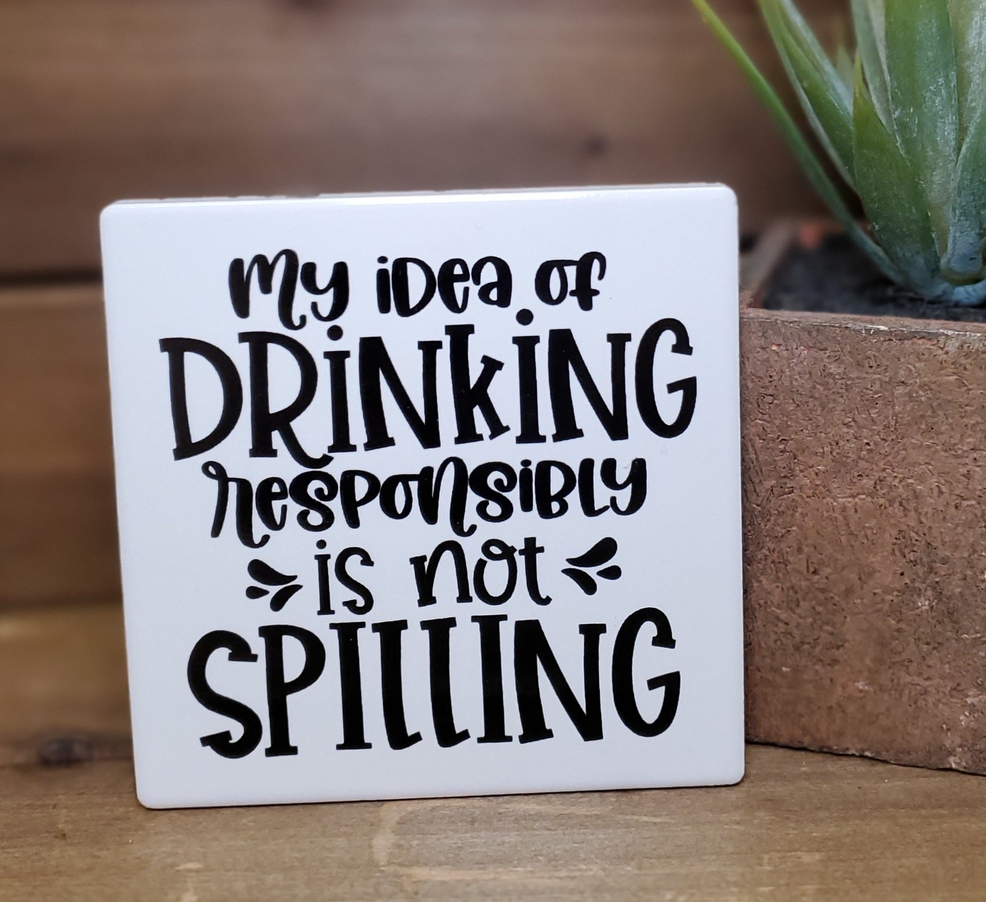 White sign with black text 'My idea of drinking responsibly is not spilling' on a wooden surface.