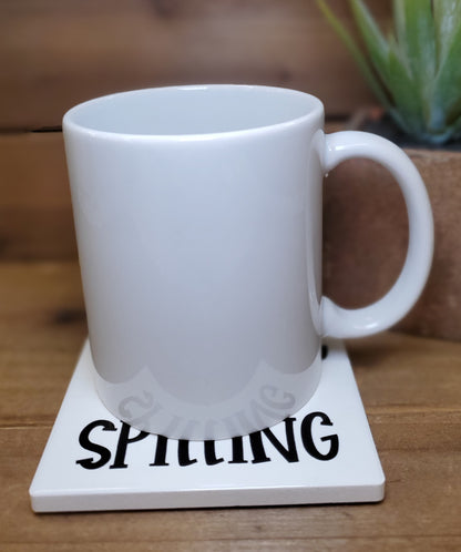 White mug on a coaster with 'SPILLING' text on a wooden surface