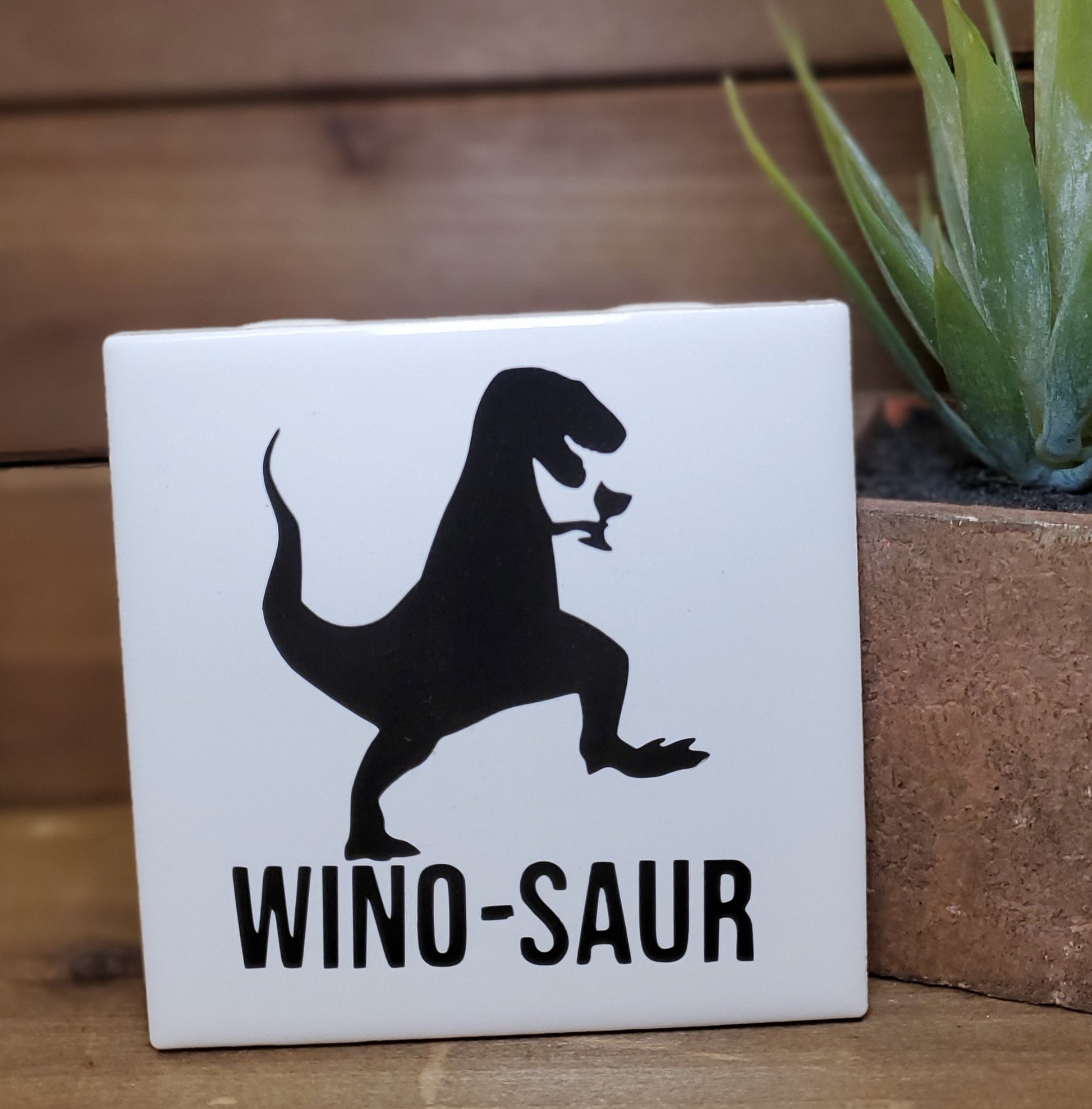 Square white tile with black dinosaur silhouette and 'WINO-SAUR' text, placed on a wooden surface with a plant in the background.