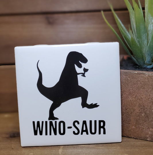 Square white tile with black dinosaur silhouette and 'WINO-SAUR' text, placed on a wooden surface with a plant in the background.