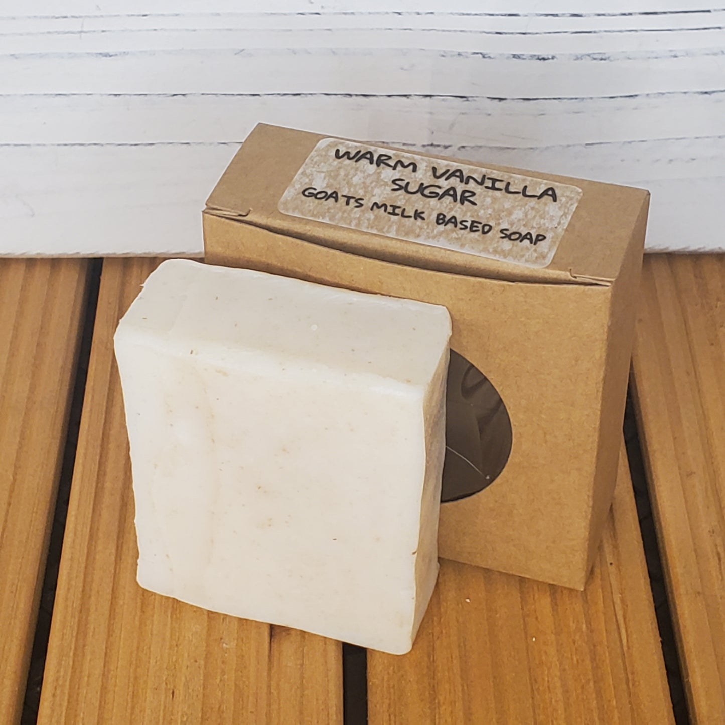 Bar of soap with a cardboard box labeled 'Warm Vanilla Sugar Goats Milk Based Soap' on a wooden surface.