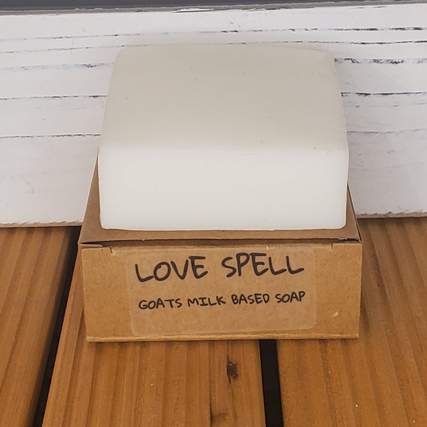 Bar of soap with a cardboard box labeled 'Love Spell Goats Milk Based Soap' on a wooden surface.