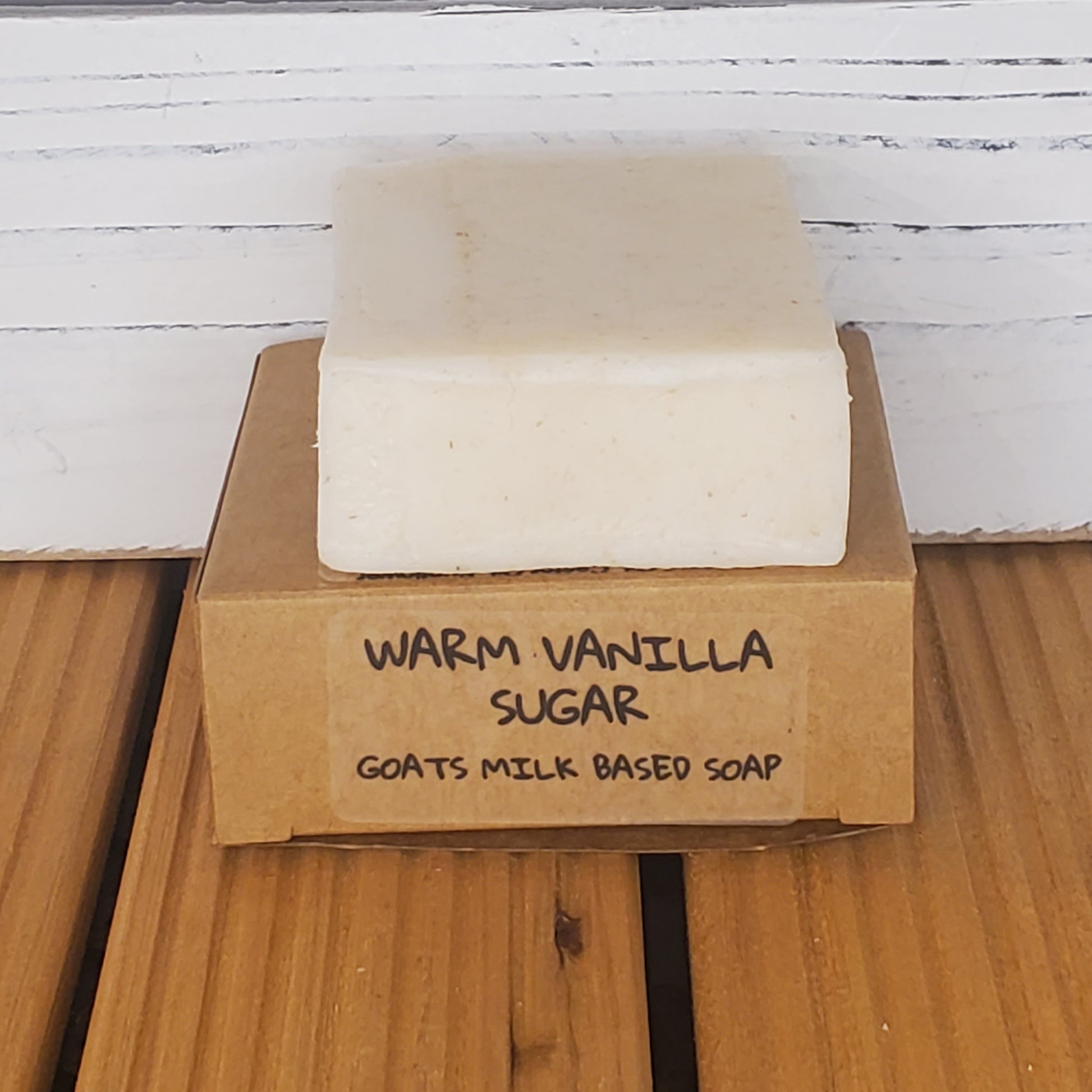 Bar of soap with a cardboard box labeled 'Warm Vanilla Sugar Goats Milk Based Soap' on a wooden surface.