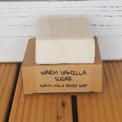 Bar of soap with a cardboard box labeled 'Warm Vanilla Sugar Goats Milk Based Soap' on a wooden surface.