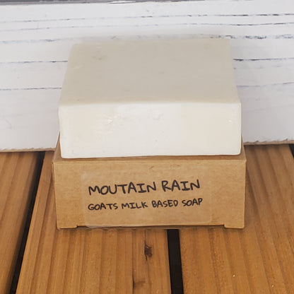 Bar of soap with a cardboard box labeled 'Mountain Rain Goats Milk Based Soap' on a wooden surface.