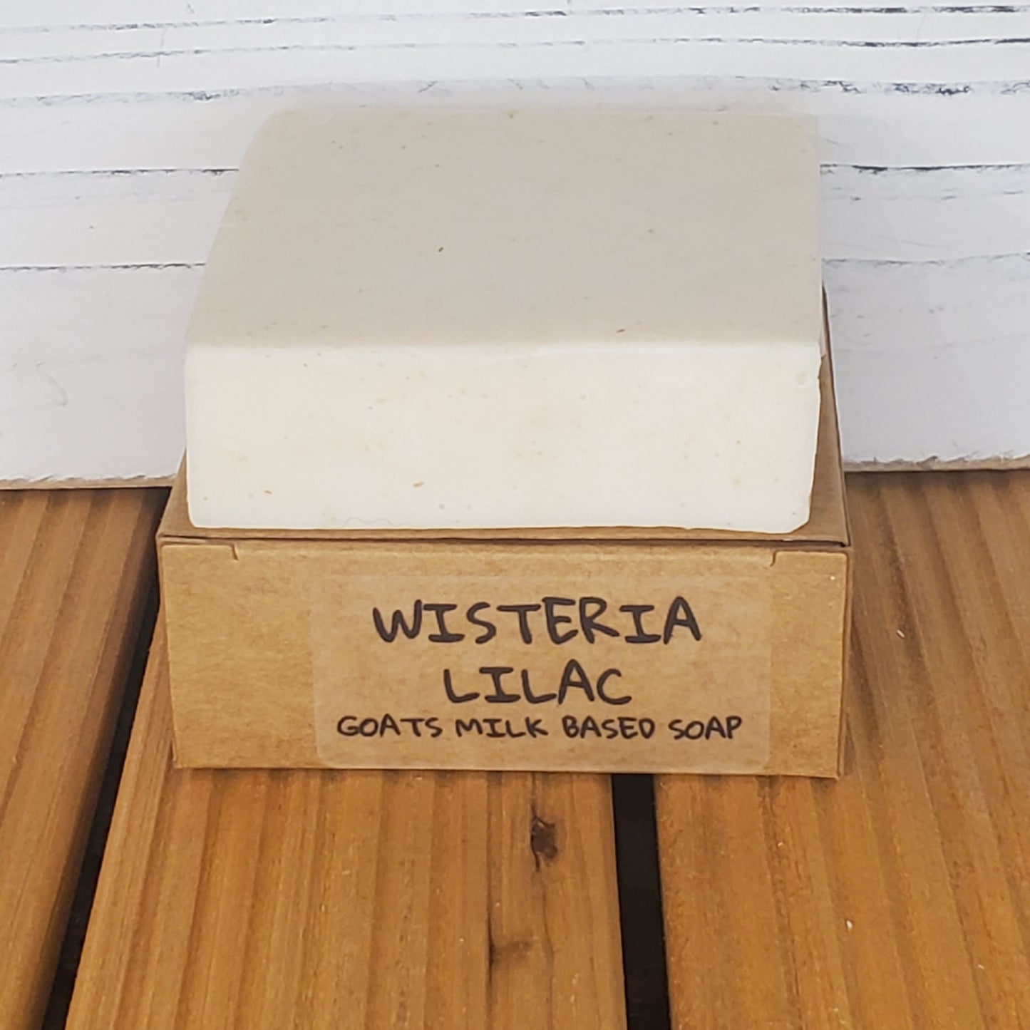 Bar of soap with a cardboard box labeled 'Wisteria Lilac Goats Milk Based Soap' on a wooden surface.