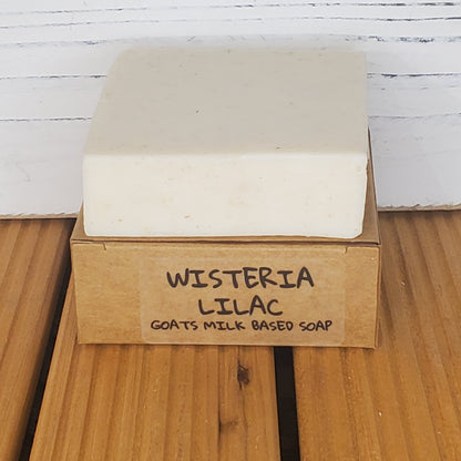 Bar of soap with a cardboard box labeled 'Wisteria Lilac Goats Milk Based Soap' on a wooden surface.