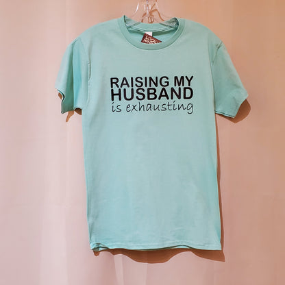 Light blue t-shirt with text 'RAISING MY HUSBAND IS EXHAUSTING' on a pink background