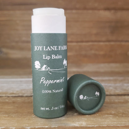 Green lip balm container with white cap on a wooden surface, featuring 'Joy Lane Farm' branding.