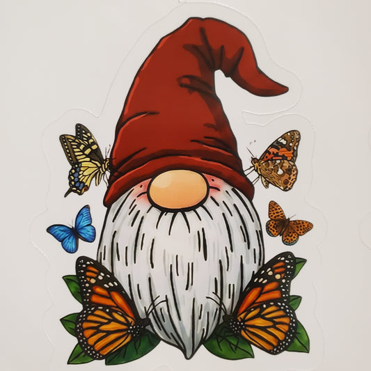 A die-cut sticker featuring a whimsical gnome with a red pointed hat, white beard, orange nose, and surrounded by orange, yellow, and blue butterflies on green leaves, shown on a plain light background.