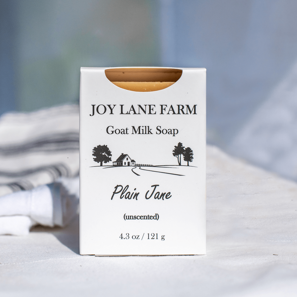 A white rectangular box of Joy Lane Farm Goat Milk Soap, labeled 'Plain Jane (unscented)', standing upright on a light fabric surface with a softly blurred background.