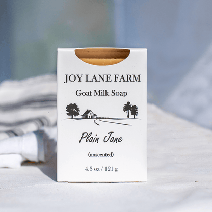 A white rectangular box of Joy Lane Farm Goat Milk Soap, labeled 'Plain Jane (unscented)', standing upright on a light fabric surface with a softly blurred background.