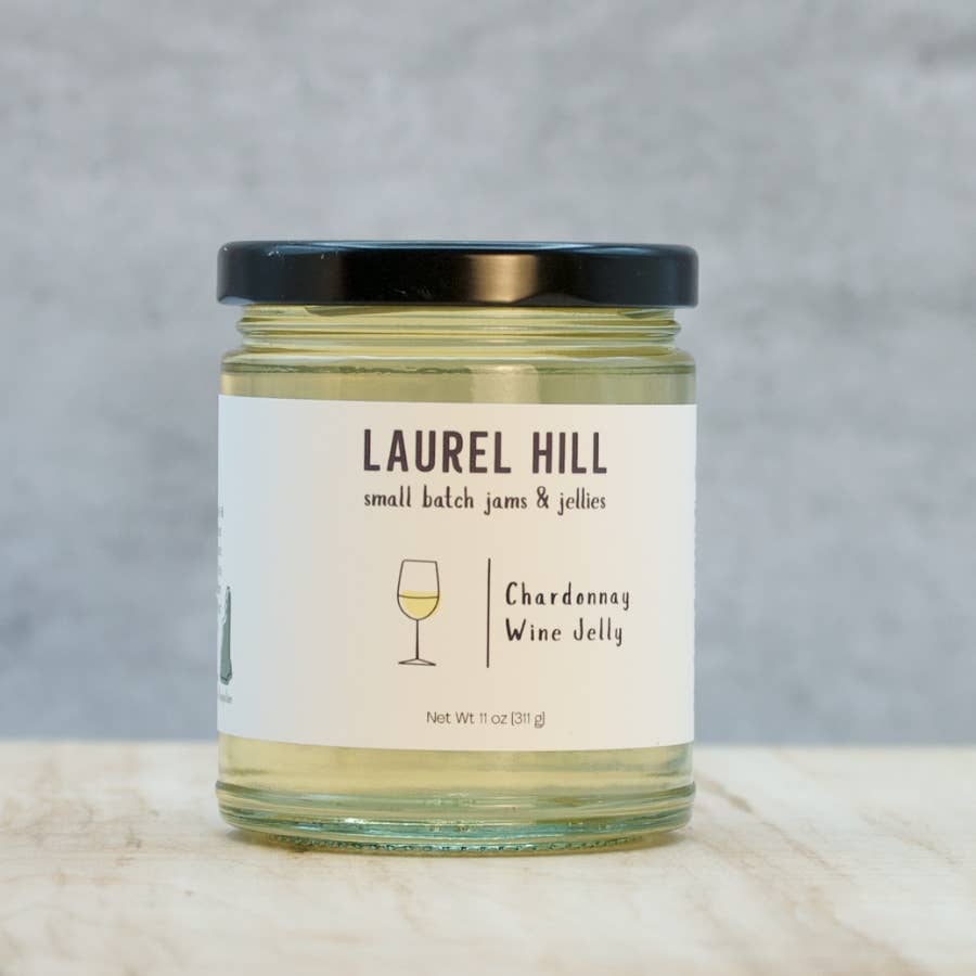 A glass jar of Laurel Hill Chardonnay Wine Jelly with a black lid and a white label, set on a light marble surface against a gray textured background.