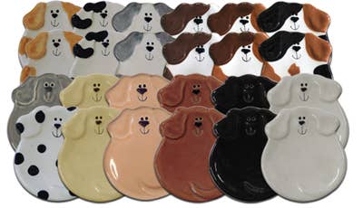A set of ceramic dog-shaped plates shown in multiple colors and patterns, including solid black, white, tan, brown, and spotted Dalmatian designs, arranged in rows against a white background.