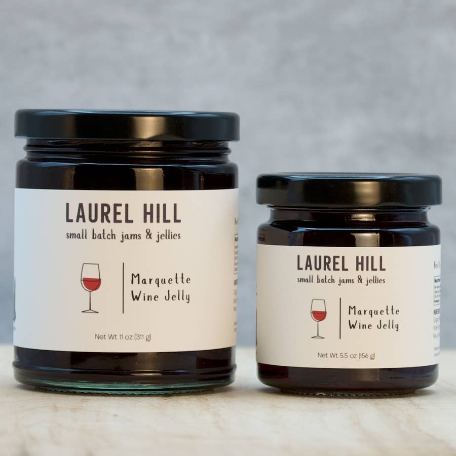 Two glass jars of Laurel Hill Small Batch Jams & Jellies, one larger jar and one smaller jar, both labeled Marquette Wine Jelly with a wine glass icon, set on a light marble surface against a textured gray background.