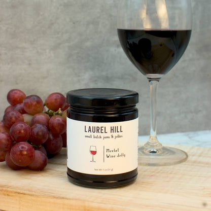 Jar of Laurel Hill Merlot Wine Jelly with a glass of red wine and grapes on a wooden surface.