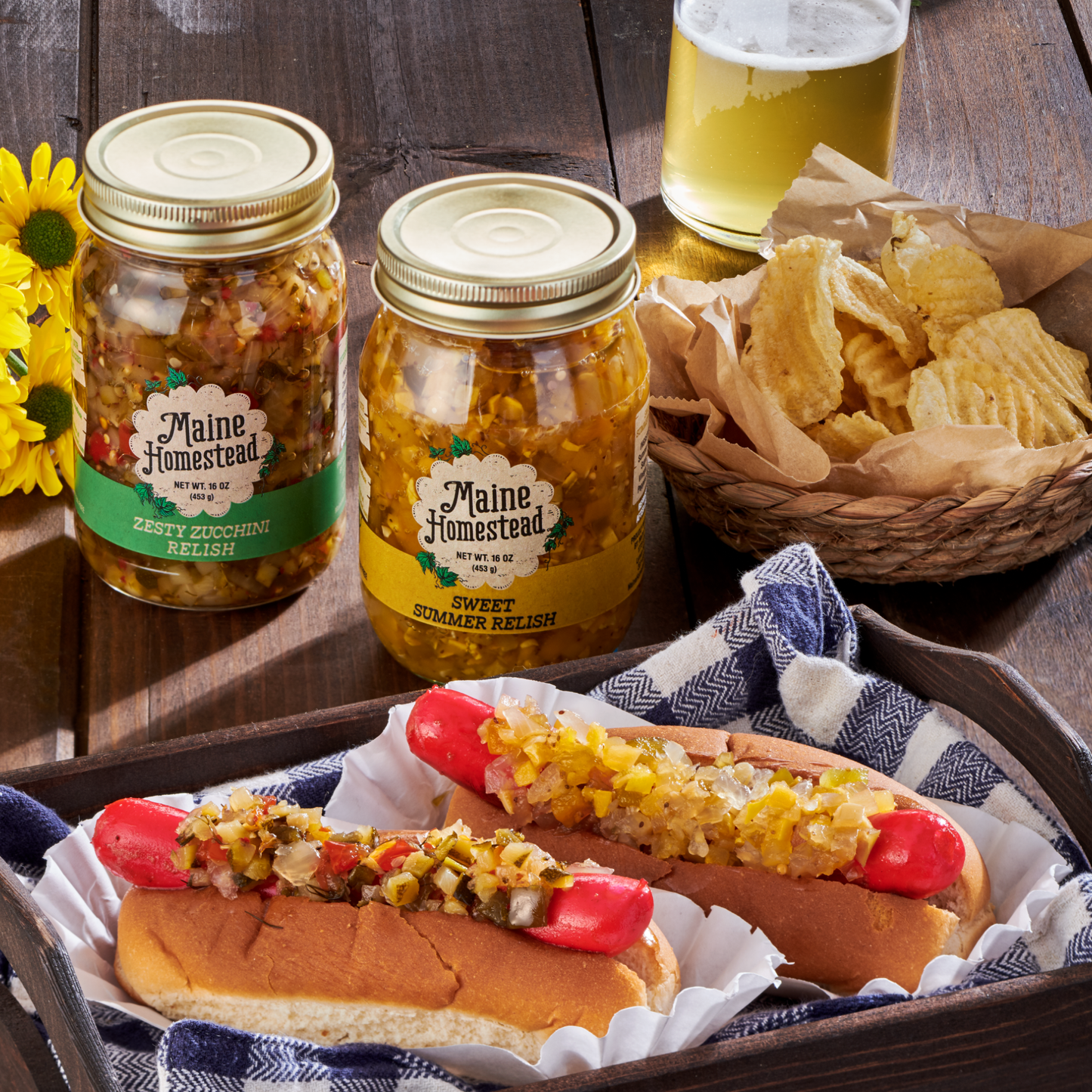 Two jars of Maine Homestead pickled relish displayed on a rustic wooden table, with two hot dogs topped with relish in the foreground and a basket of potato chips and a glass of beer nearby.